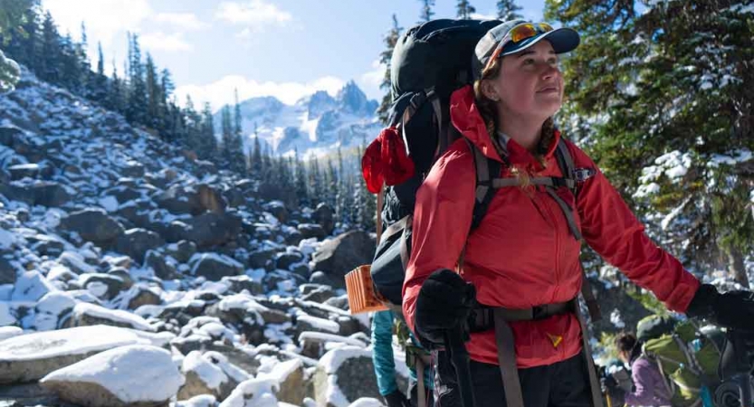 a person carrying a backpack stands on a rocky landscape and smiles contentedly. there are mountains in the background.